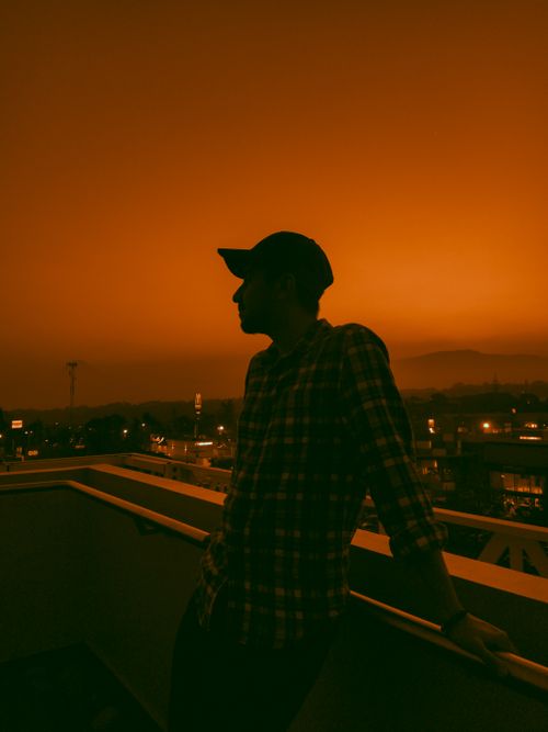 Free A man in a baseball cap leans on a rooftop railing, silhouetted against an orange sunset in an urban setting. Stock Photo