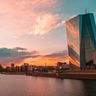 The glass facade of the European Central Bank headquarters in Frankfurt reflects a warm, vibrant sunset.