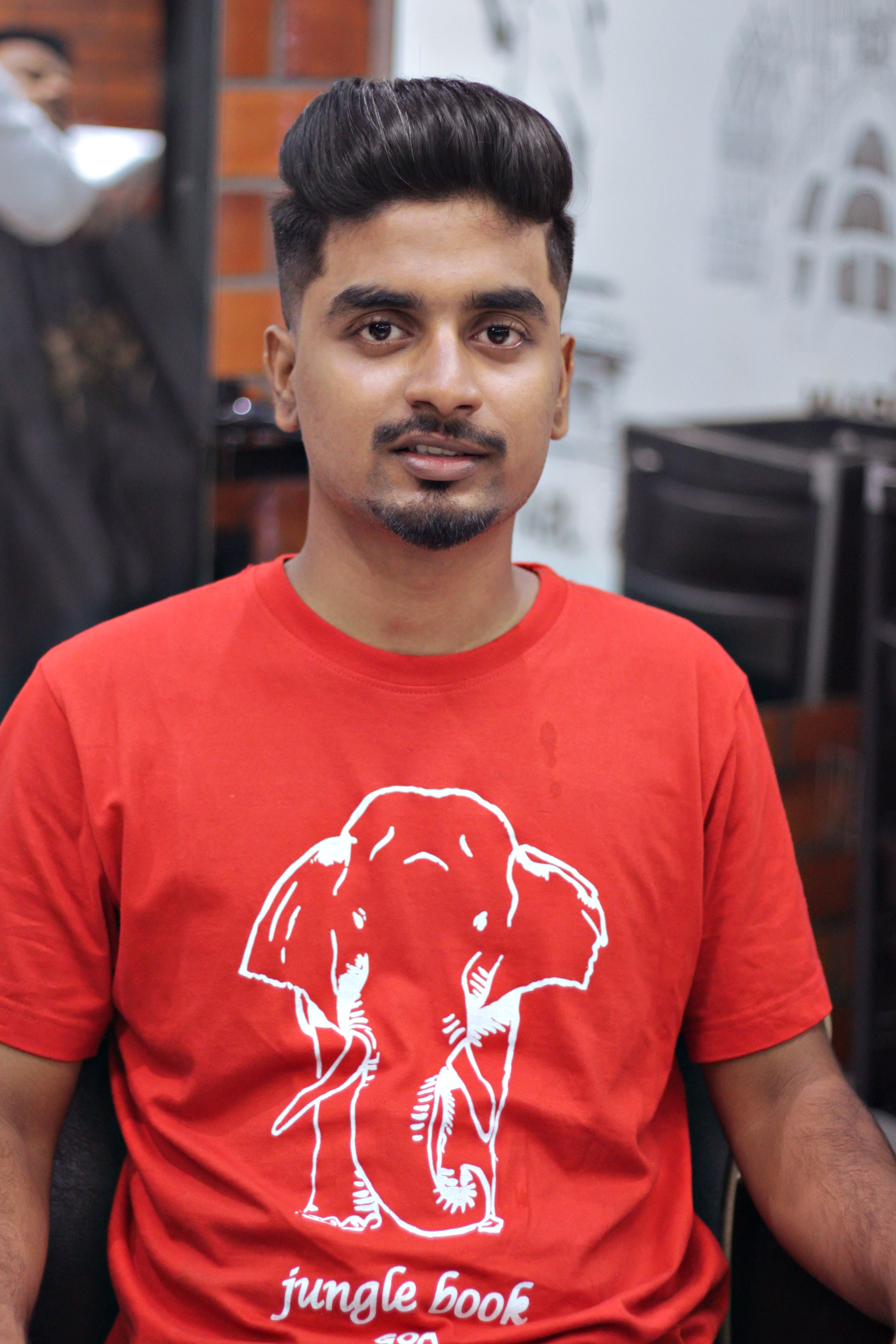 Portrait of a young man in a red graphic t-shirt seated indoors with a modern hairstyle.