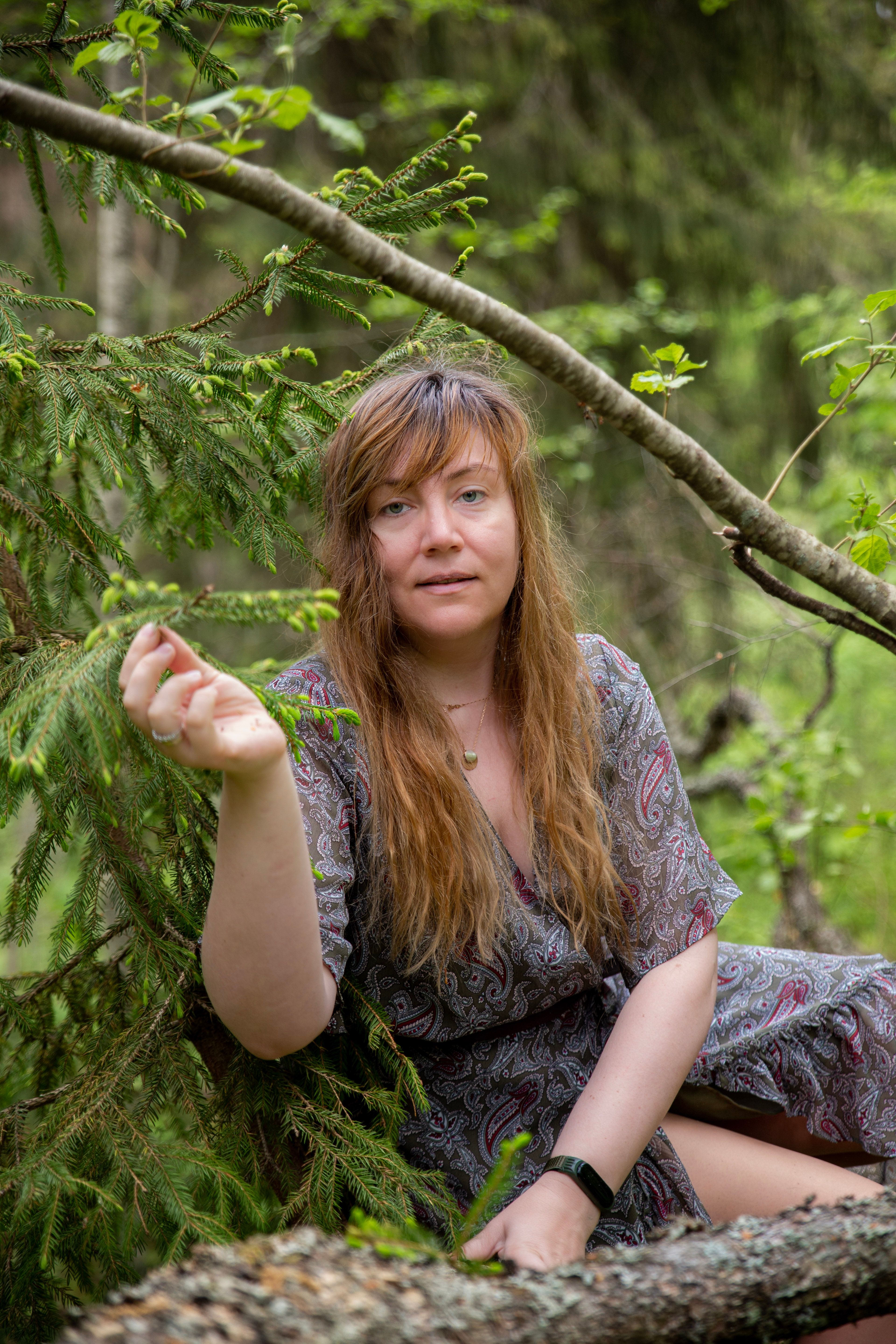 A woman in patterned dress sitting amidst coniferous trees in a serene forest setting.