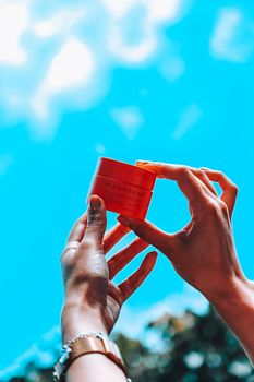 Hands reaching towards a vibrant blue sky, holding a skincare jar symbolizing beauty and care.