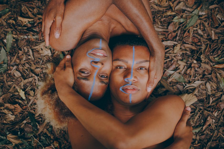 Creative outdoor portrait of two shirtless men with artistic blue face painting, lying on leaves.