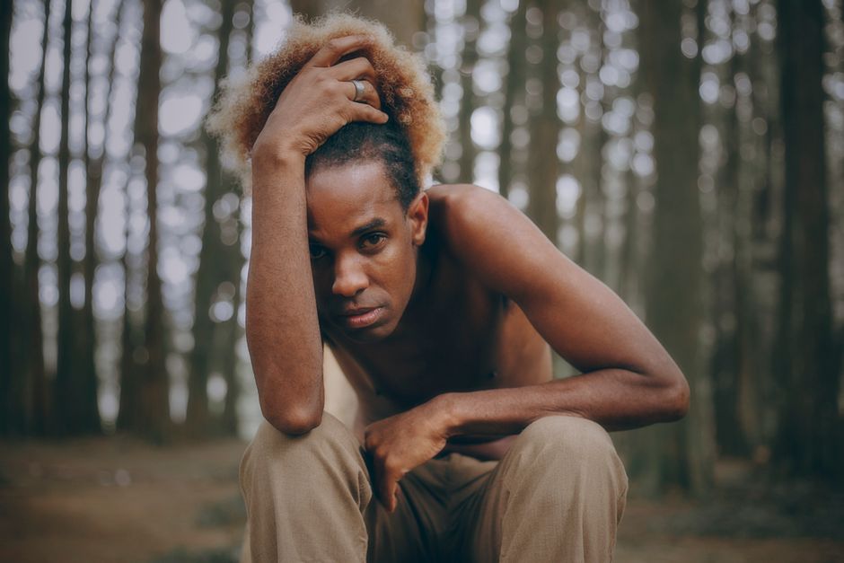 A shirtless man with afro hairstyle poses thoughtfully in a wooded area.