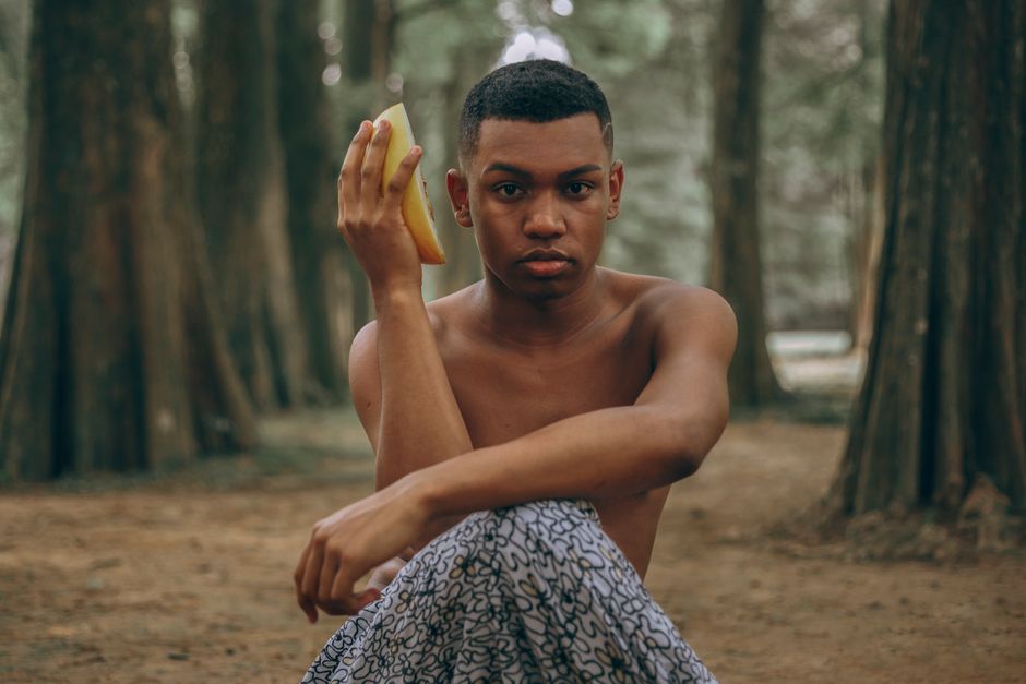A young man sitting outdoors in a forest holding a banana with a thoughtful expression.