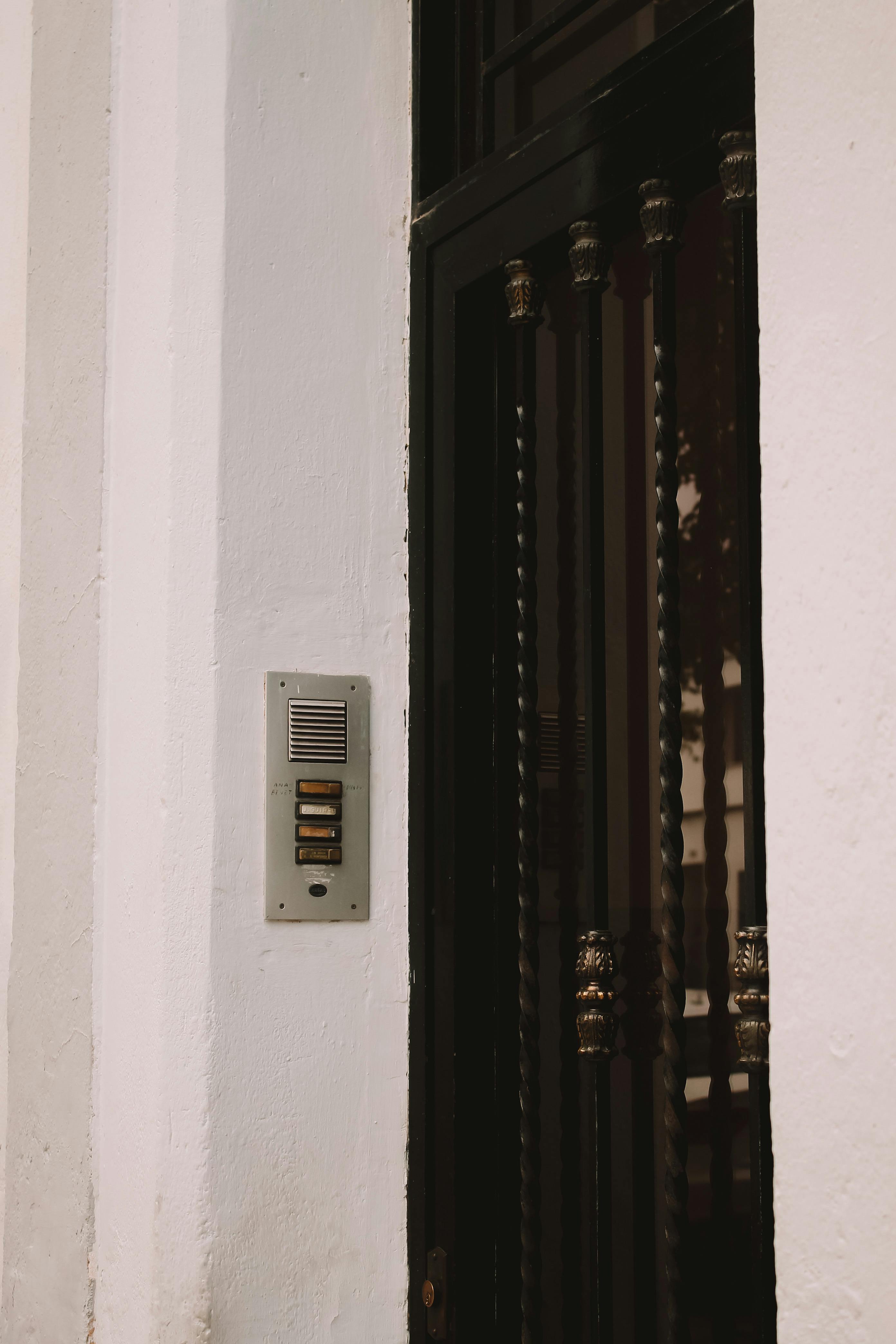 Detailed view of an intercom system on the exterior wall of a residential building, featuring a black iron door.