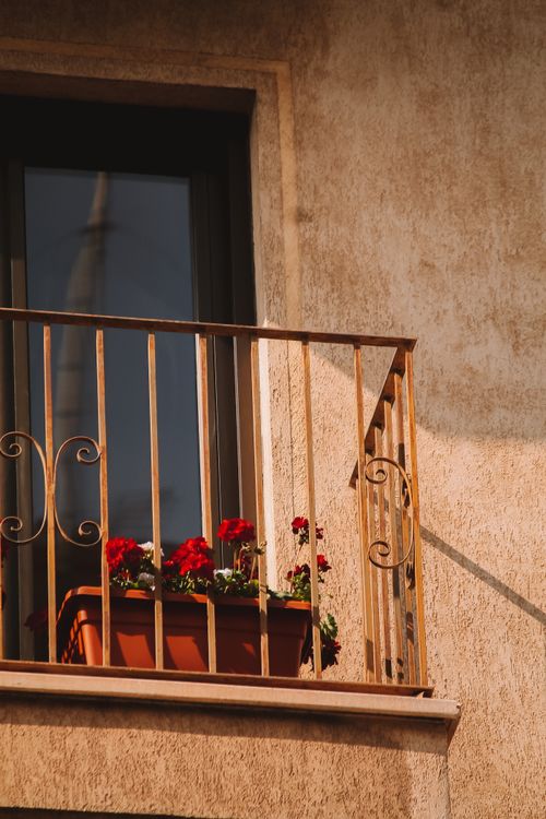Free A sunlit balcony adorned with vibrant red flowers in an urban setting, showcasing architectural elegance. Stock Photo