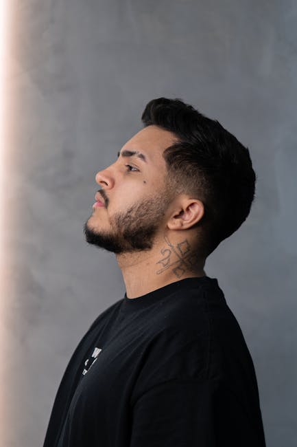 Young man with beard and neck tattoo posing against a grey background in side profile view.