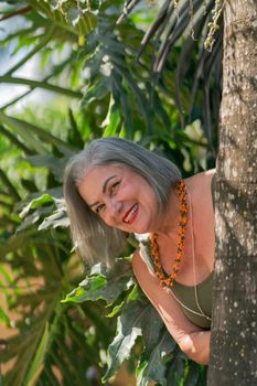 A joyful senior woman with gray hair smiles while surrounded by lush tropical greenery.