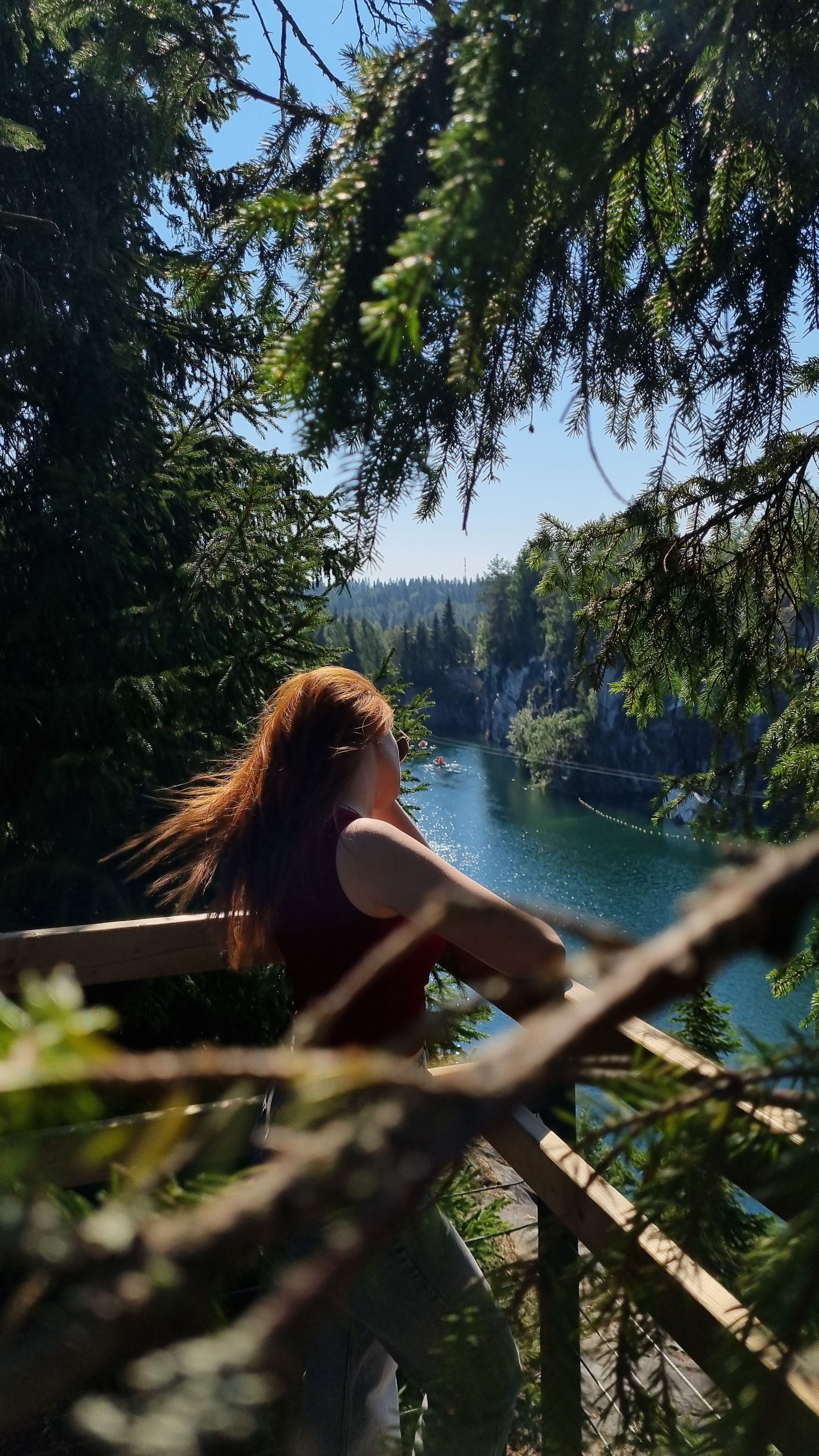 A woman stands on a forest balcony overlooking a tranquil lake with clear skies.