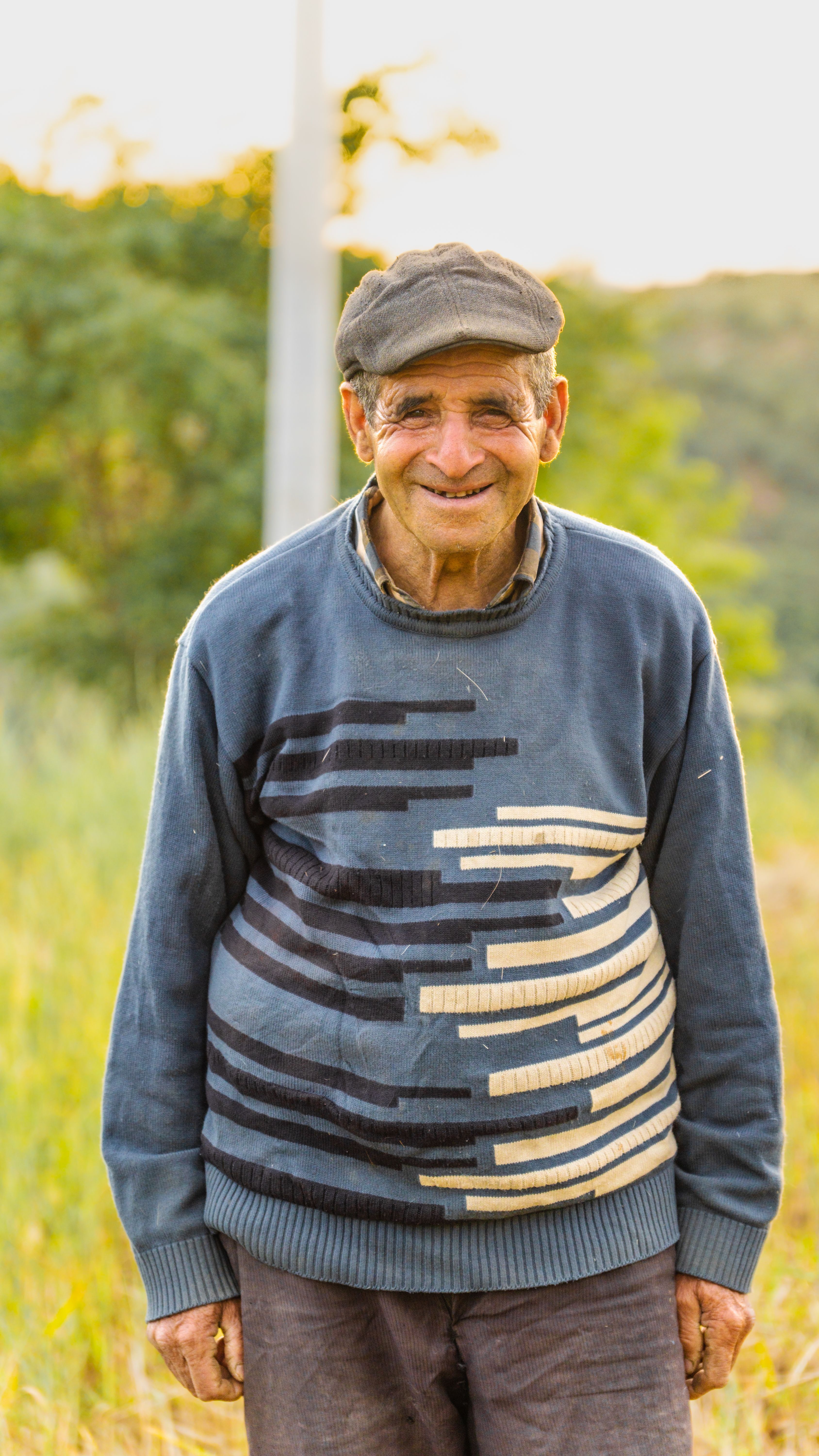 Portrait of an elderly man with a cap smiling in a rural outdoor setting.