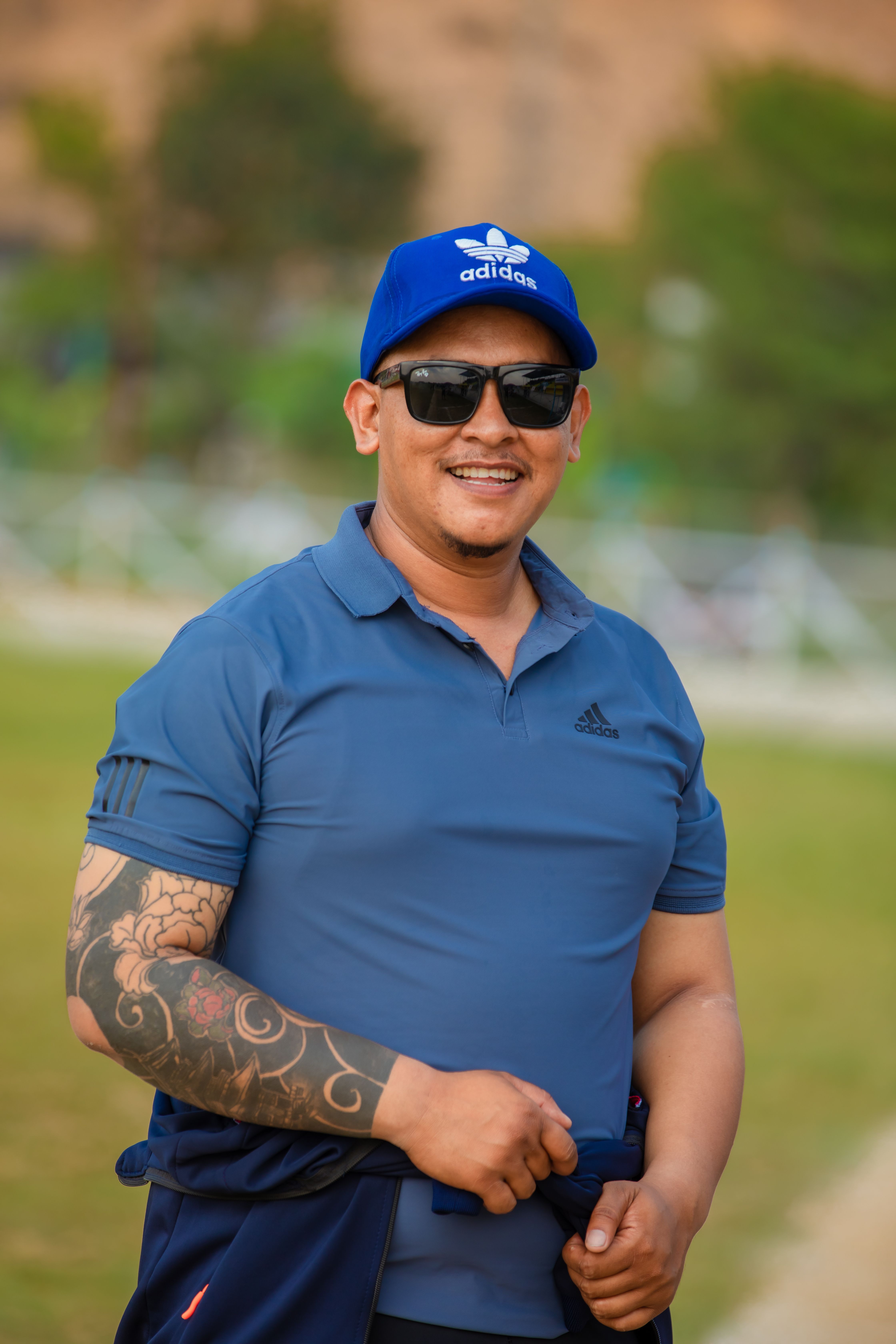 A cheerful man poses in casual blue attire, showcasing tattoos and sunglasses, in an outdoor setting.