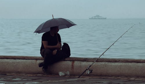 A Man Sitting on the Pier, Holding an Umbrella and Fishing