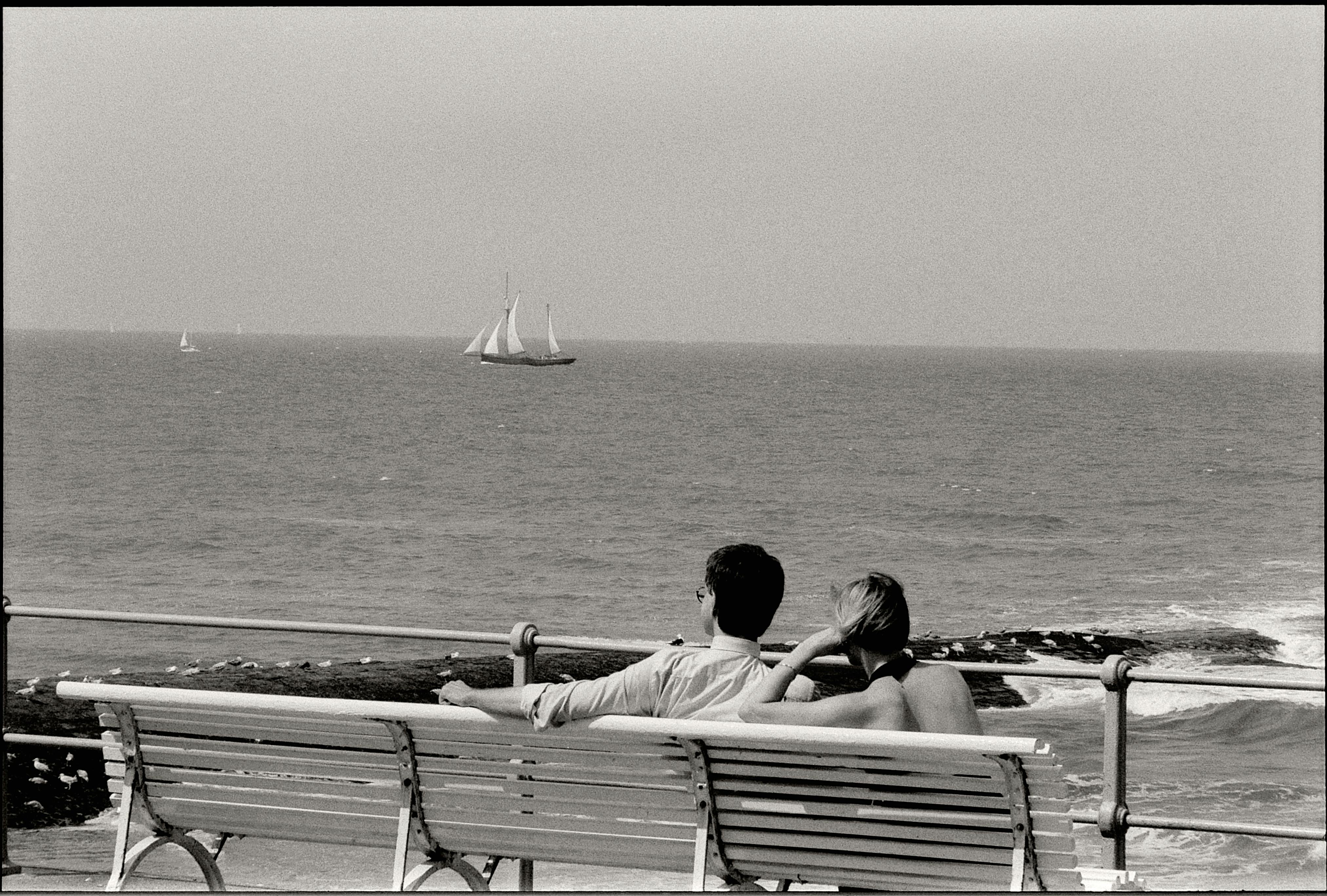 A couple enjoys a peaceful seaside view, sitting on a bench in black and white.