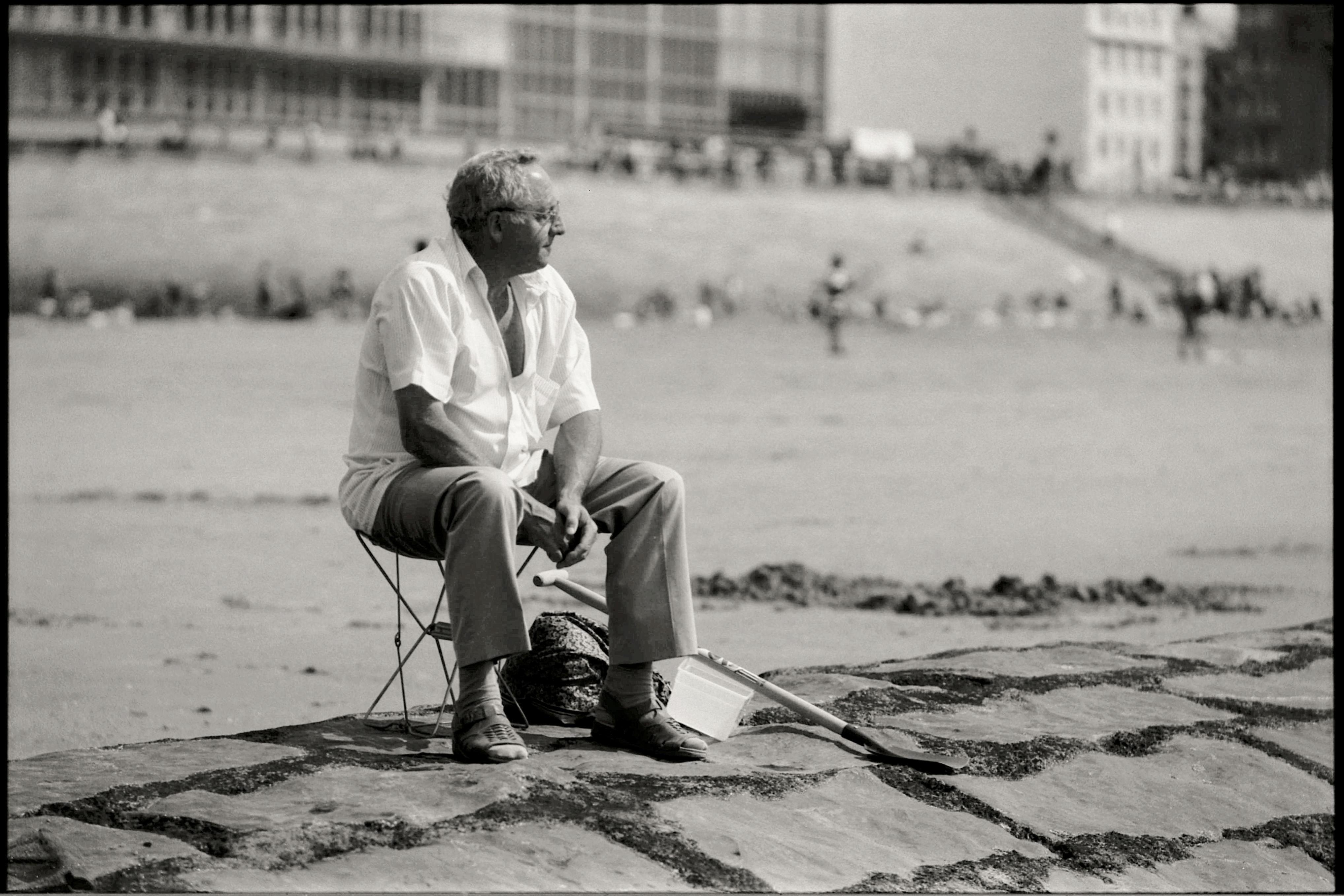Elderly man sits on a folding chair at the beach, enjoying quiet solitude in a vintage black and white photograph.