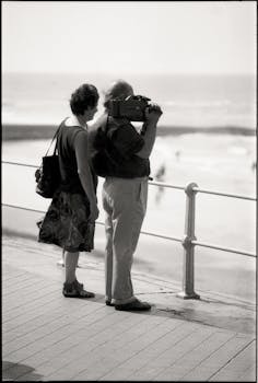 A couple stands on a promenade by the sea, capturing the view with a vintage camera.