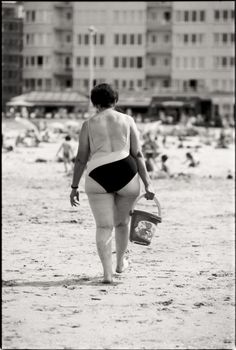 Black and white photo of a woman holding a bucket walking on a sandy beach.