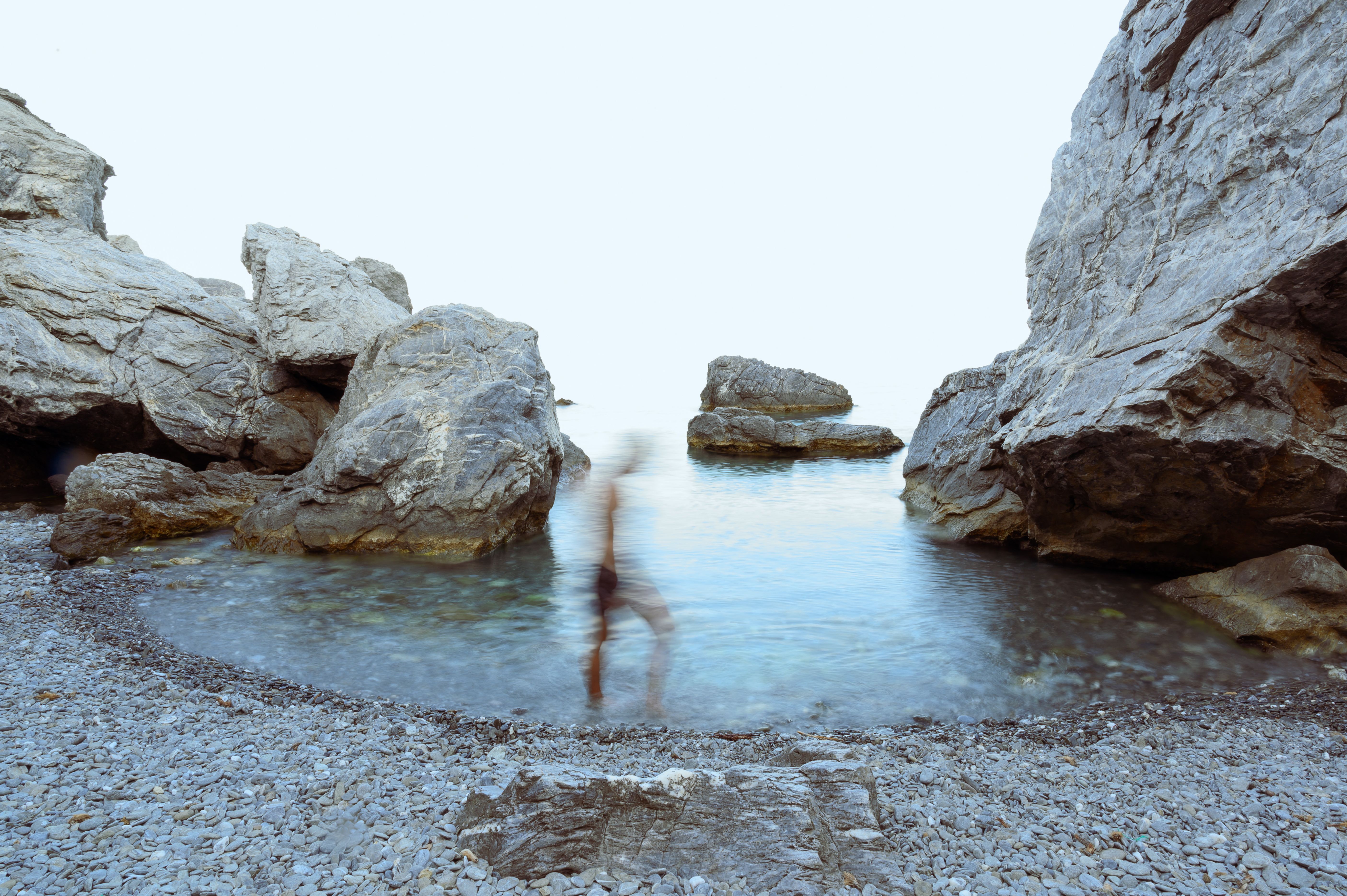 A serene rocky shore with a blurred figure in the water at sunrise, blending art and nature.