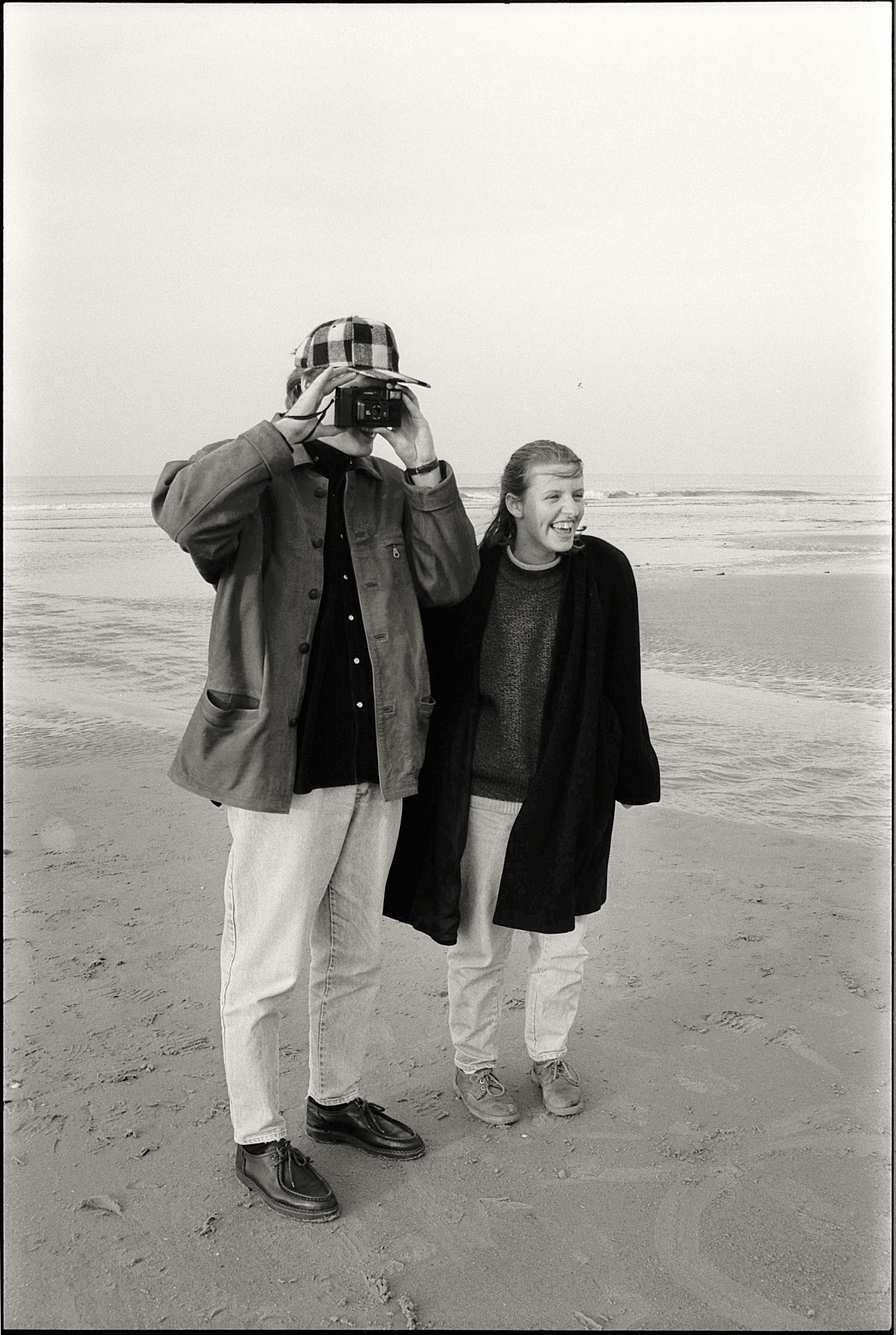 Black and white image of a couple on a beach, capturing the moment with a camera.
