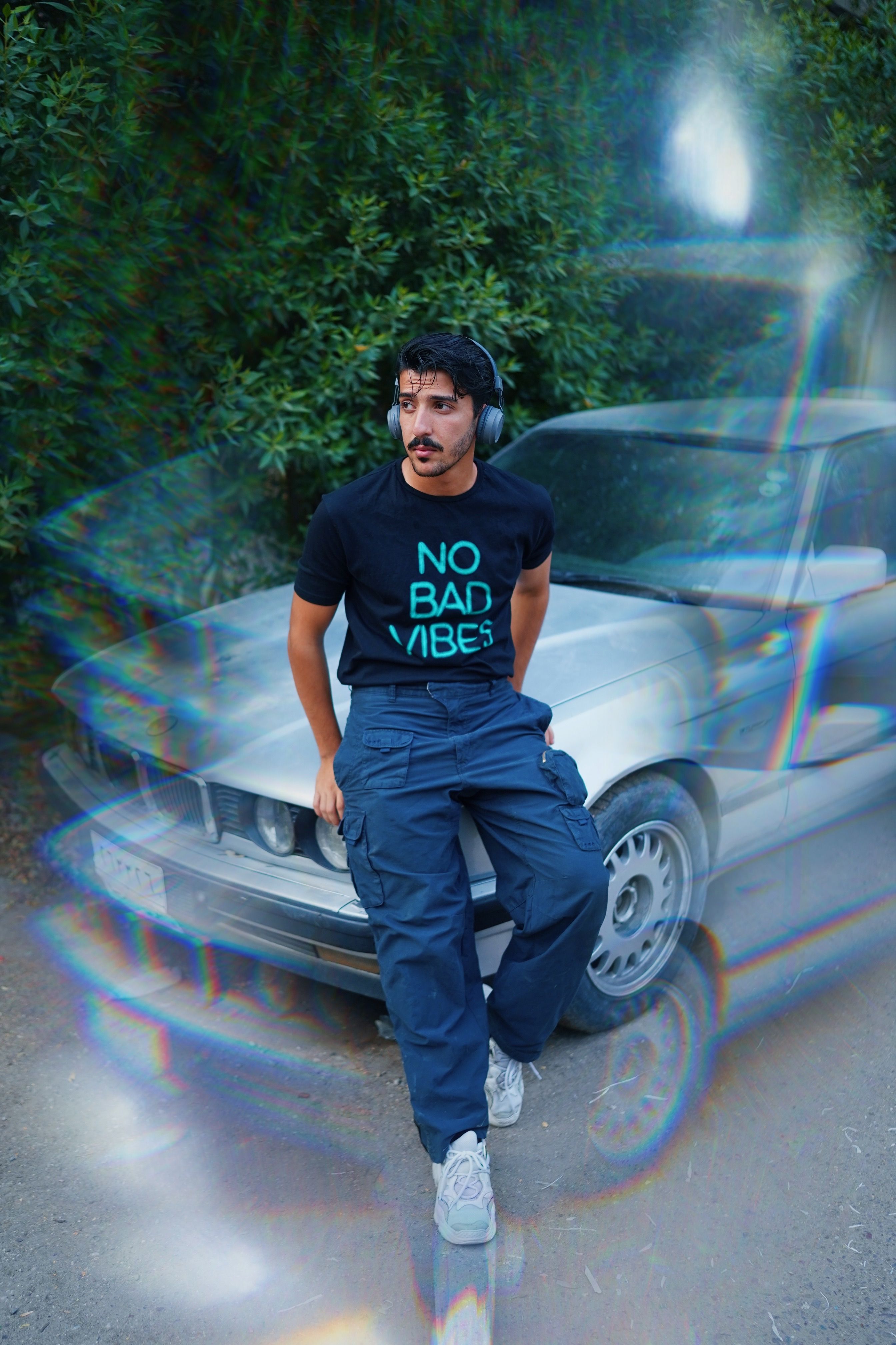 Young man in casual attire with headphones, leaning on a car outdoors.