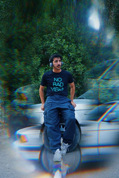 Free Young man wearing headphones and streetwear, relaxing against a car on a vibrant day. Stock Photo