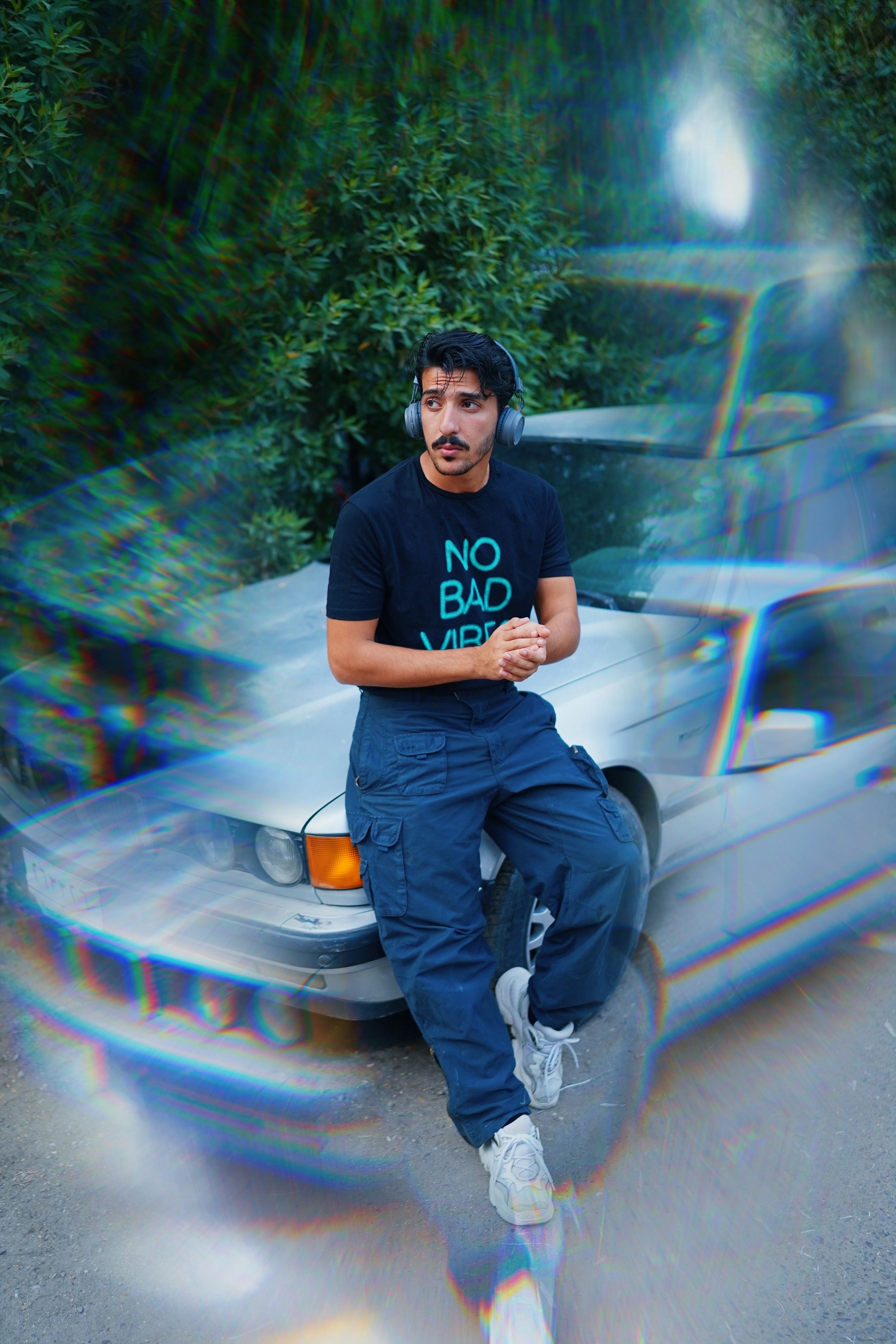 A man in casual streetwear and headphones leans on a vintage car surrounded by nature, exuding urban vibes.