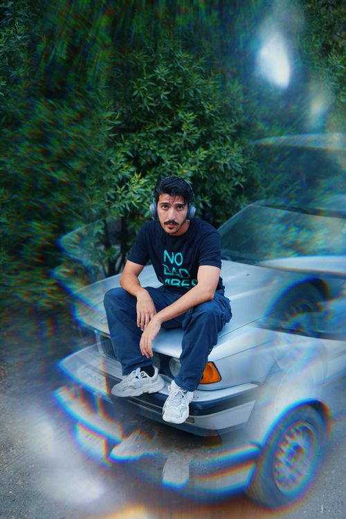 Free A man in casual clothing sits on a car hood, wearing headphones with trees in the background. Stock Photo