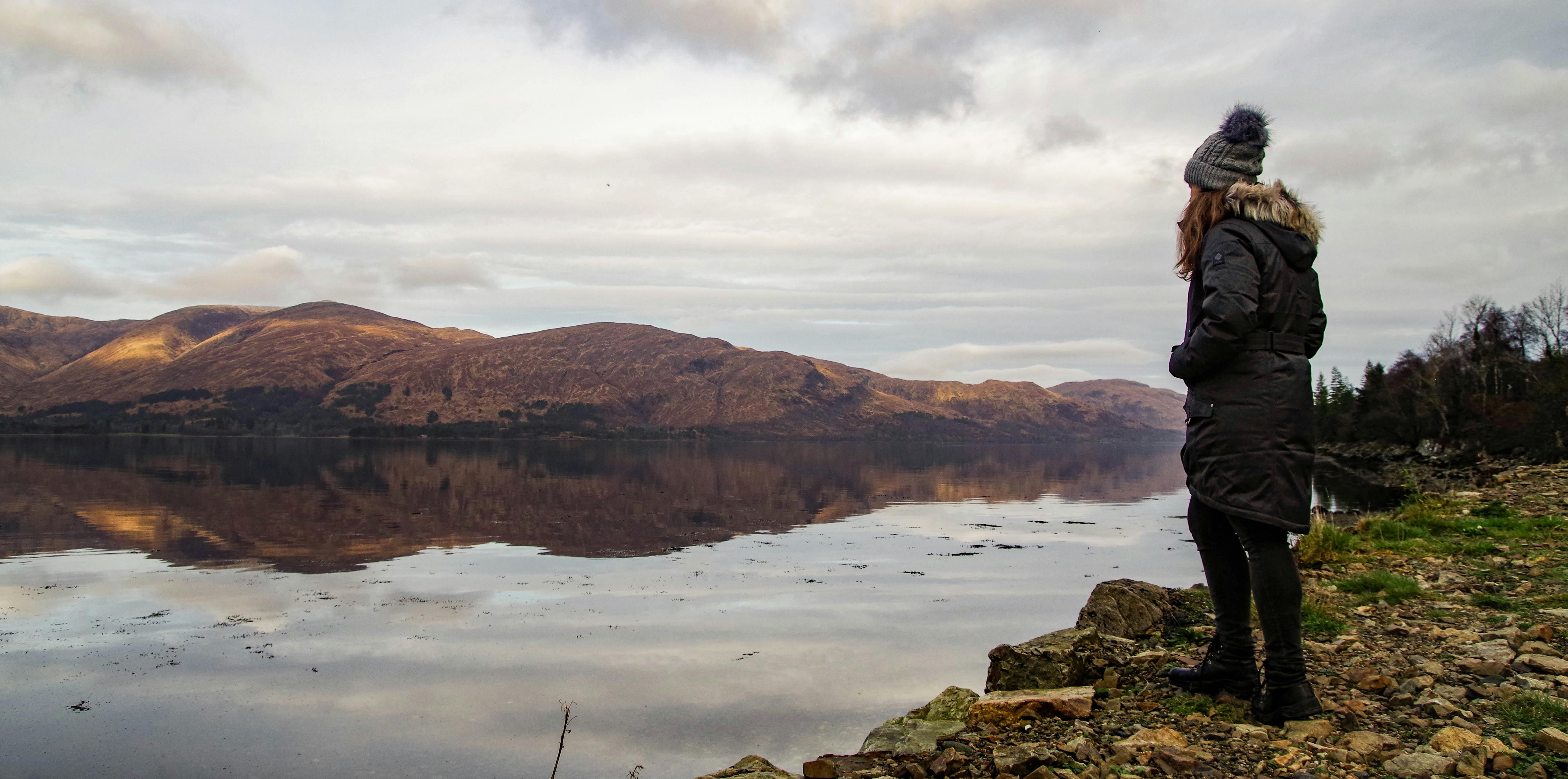 一名穿着冬装的妇女凝视着平静的湖泊，在多云的天空下，湖面倒映着周围的山脉