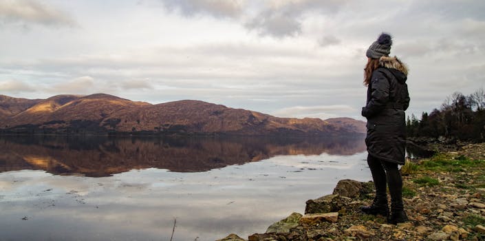 一名穿着冬装的妇女凝视着平静的湖泊，在多云的天空下，湖面倒映着周围的山脉