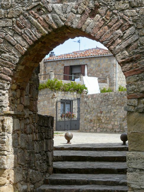 Fotomural Un arco de piedra rústico enmarcando un patio iluminado por el sol con una casa tradicional.