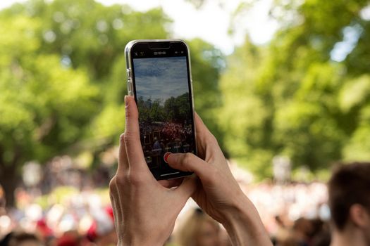 Hands holding a smartphone, recording an outdoor event with bright greenery.
