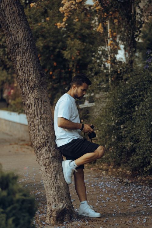 Free A young man in casual attire leans against a tree in a park, holding a camera and enjoying the outdoor ambiance. Stock Photo