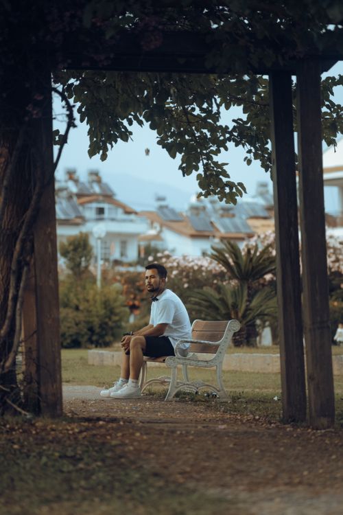 Free A man in a white t-shirt sits on a park bench amidst trees and urban scenery. Stock Photo
