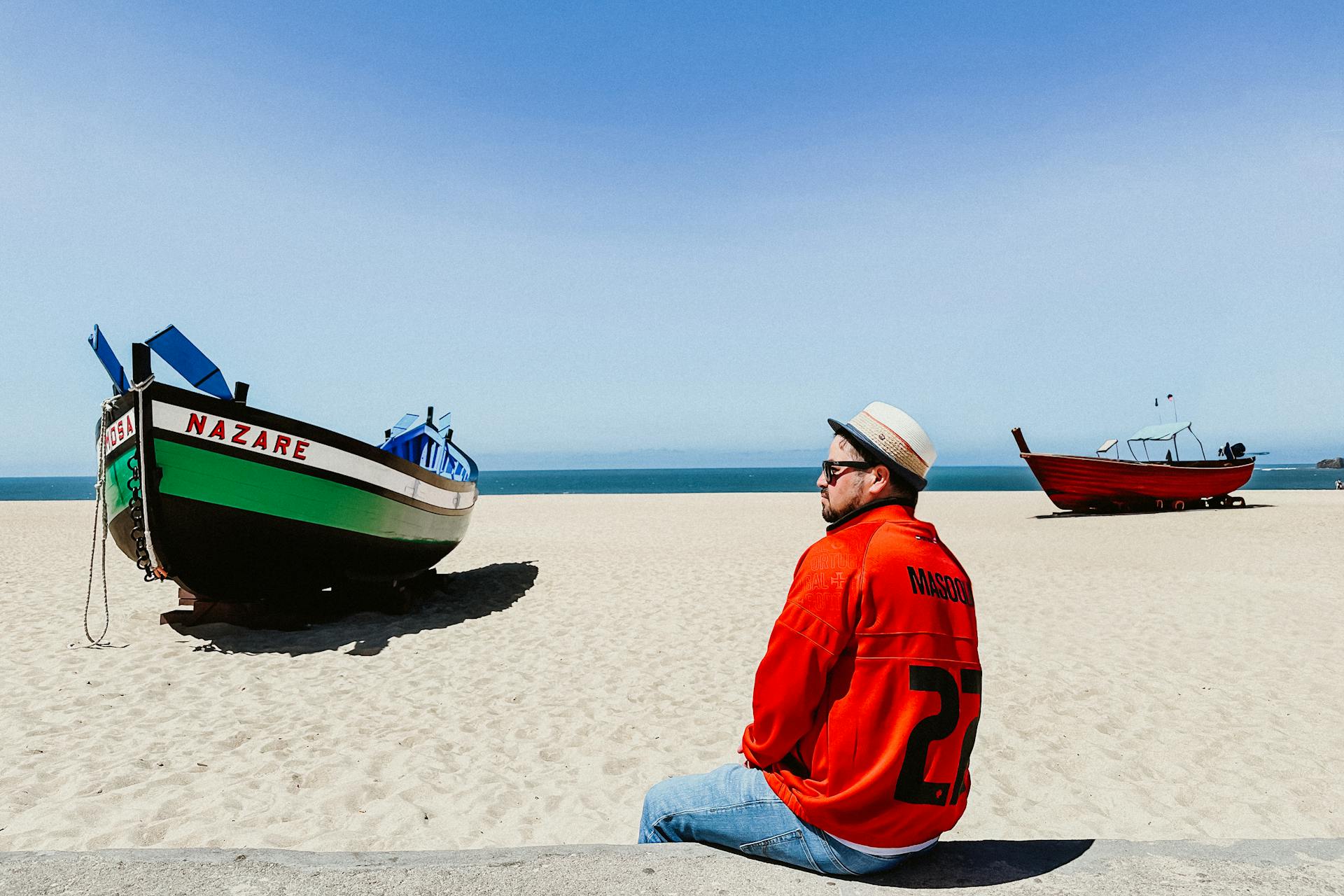A man in a red jersey relaxes on Nazare beach, Portugal, next to colorful traditional fishing boats under clear blue skies.