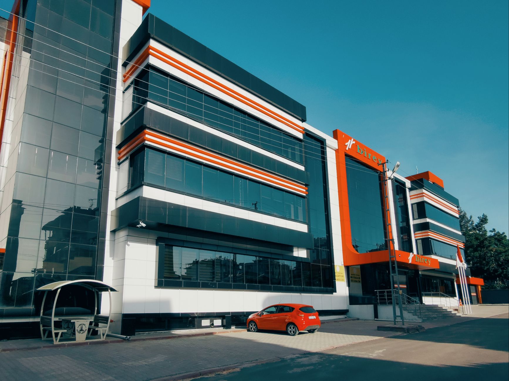 Contemporary building with glass facade and vibrant red accents, beside a parked car under clear blue sky.