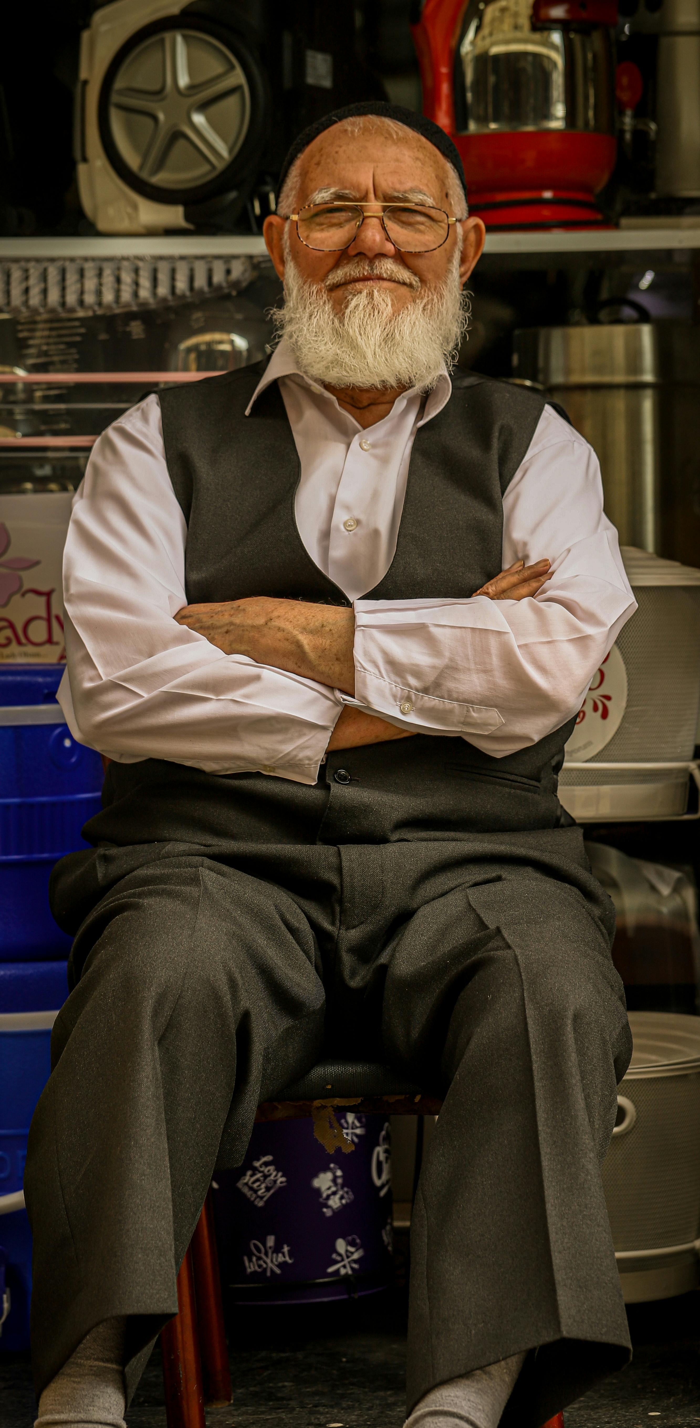 A senior man with a white beard, wearing a vest and suit, sits with arms crossed in an indoor setting.