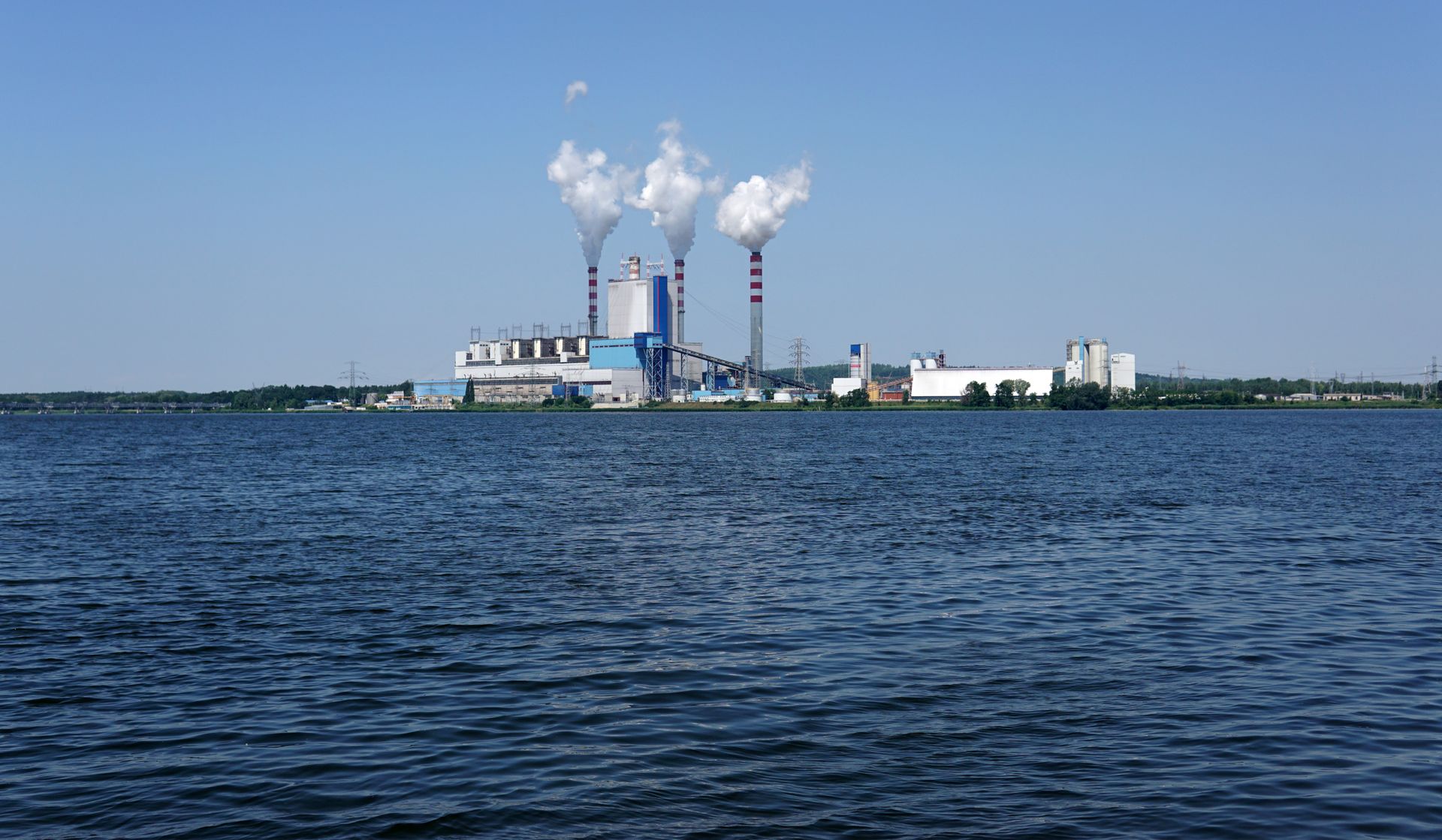 A power plant with smoke stacks by a calm lake under a clear sky.