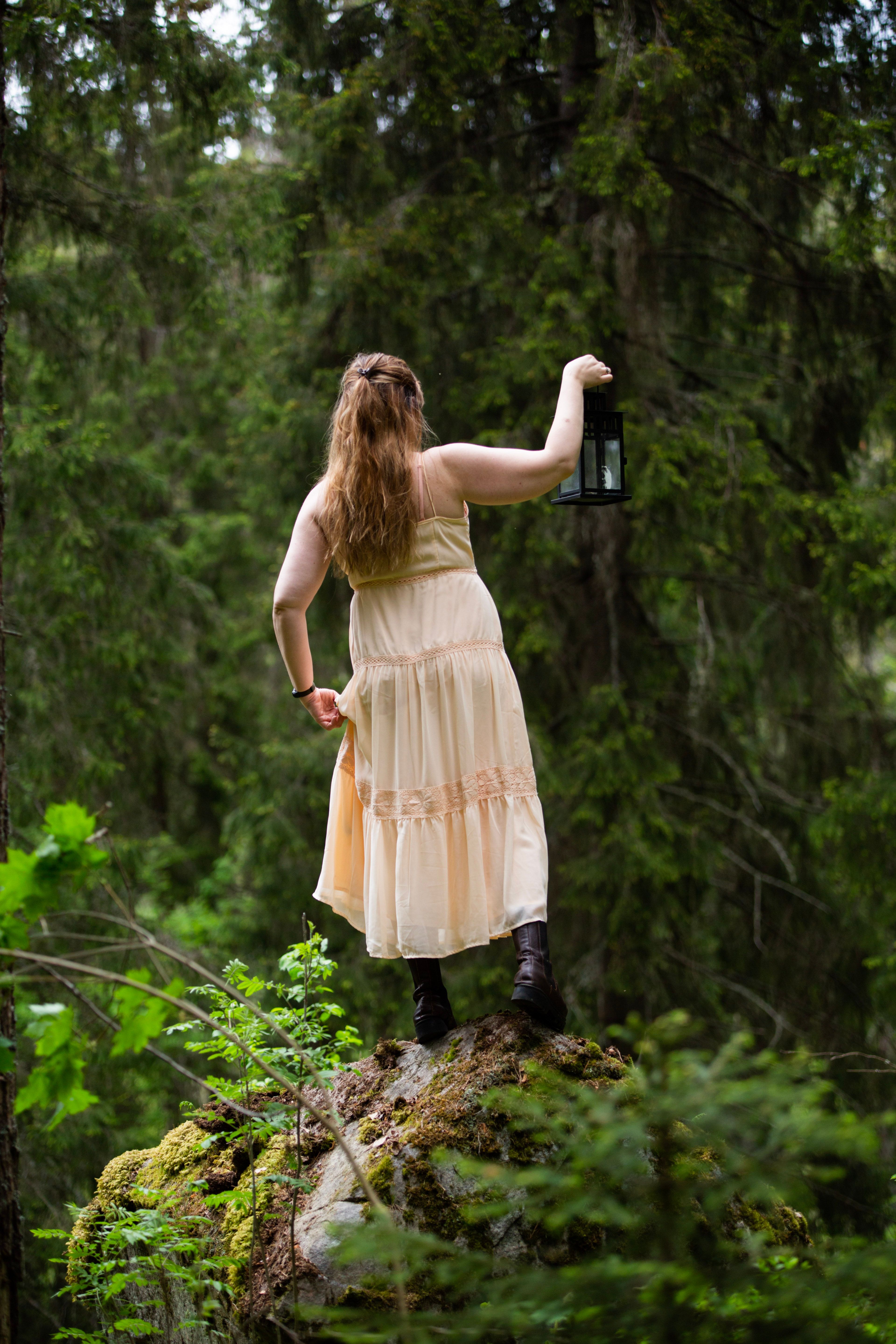 A woman in a flowing dress stands in a lush forest holding a lantern, creating a mysterious ambiance.