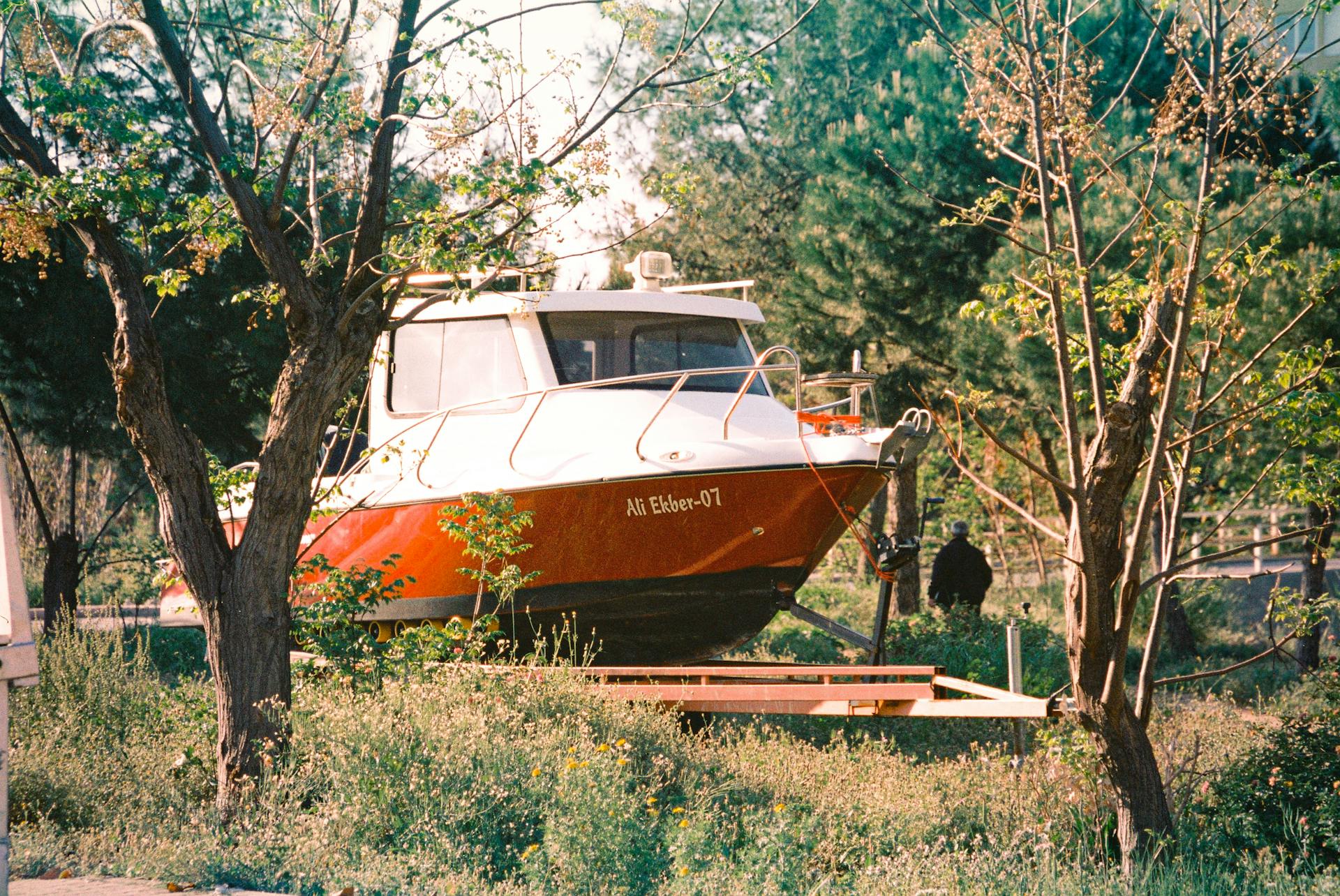A vibrant red boat sits on a trailer amidst lush greenery and tall trees in a peaceful rural setting.