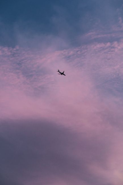 A lone airplane flies against a purple sky, highlighted by soft twilight clouds, creating a serene scene.