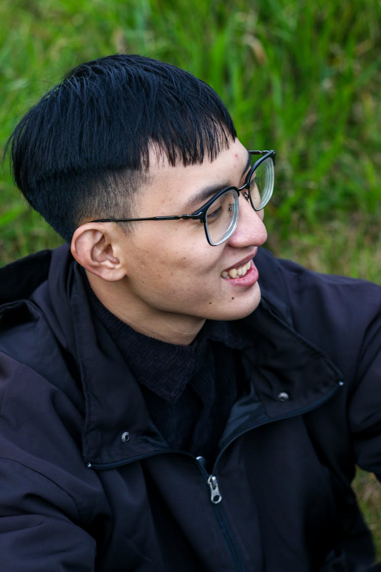 Young Man In Eyeglasses And A Black Jacket Sitting Outside On The Grass