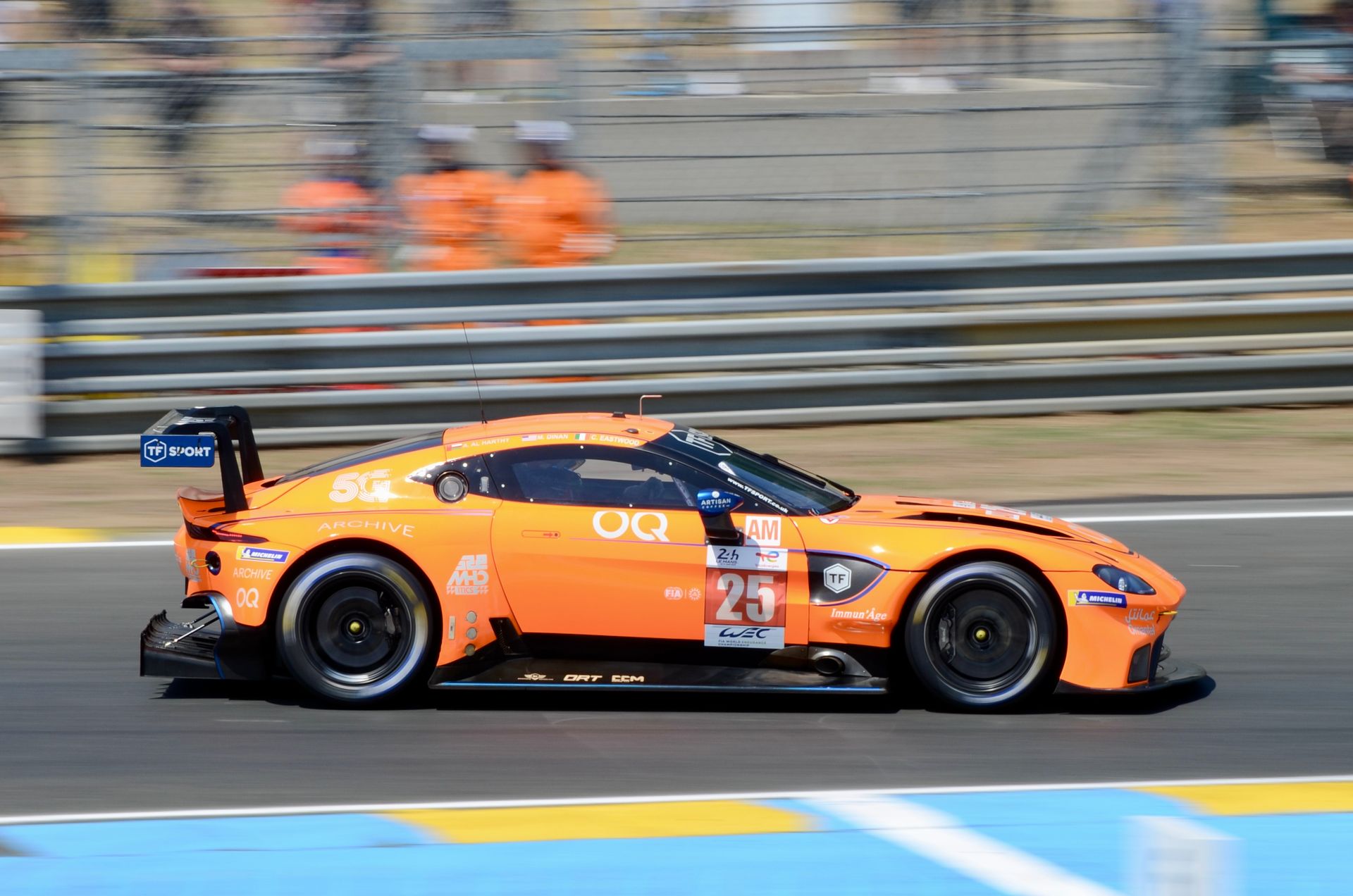 A bright orange Aston Martin on the Le Mans circuit, captured during a motorsport endurance race.