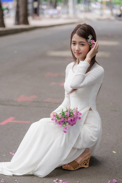 Woman in White Dress Crouching on Street with Flowers in Hands