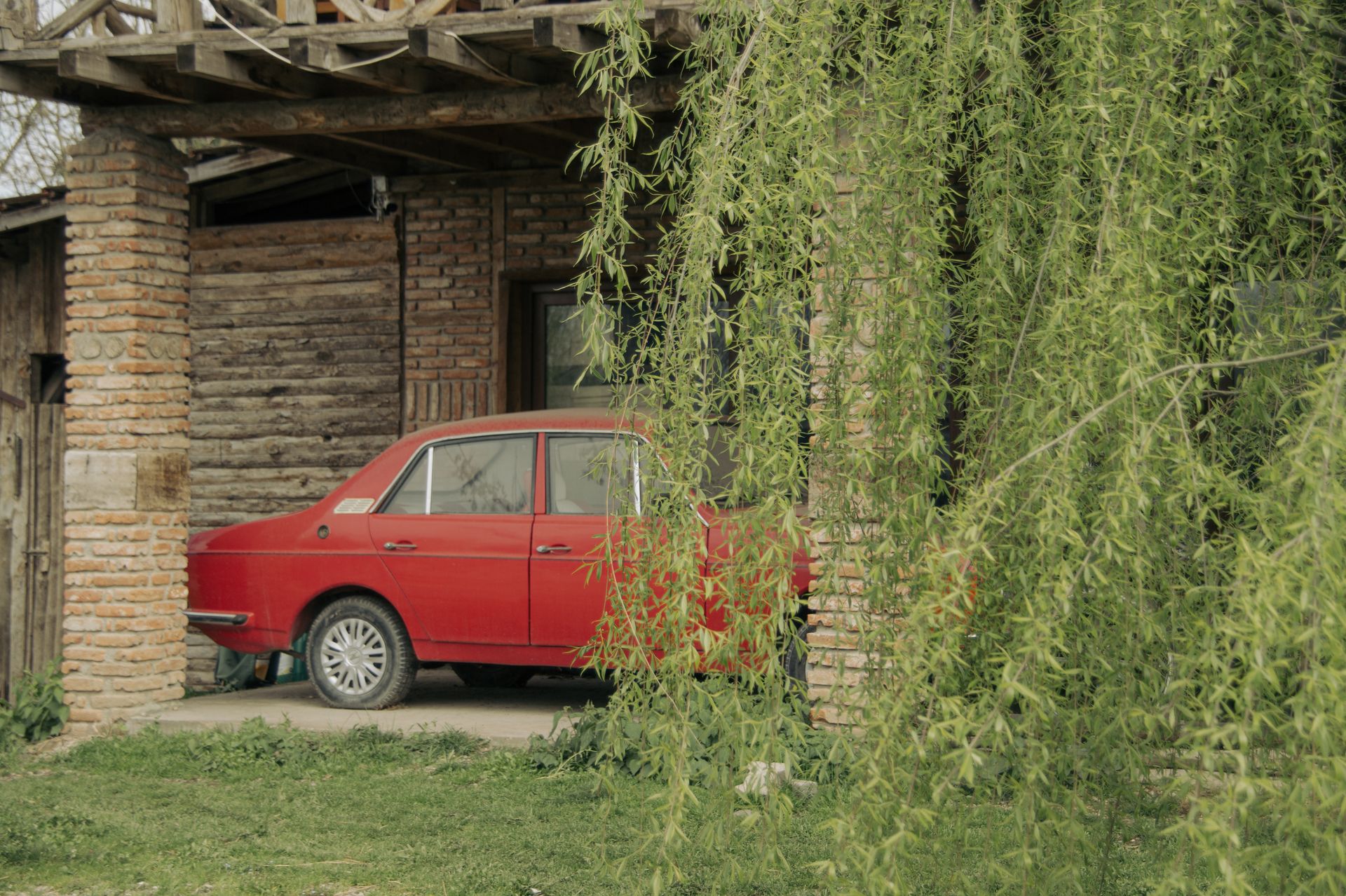 A classic red car parked under the balcony of a rustic brick house with greenery.