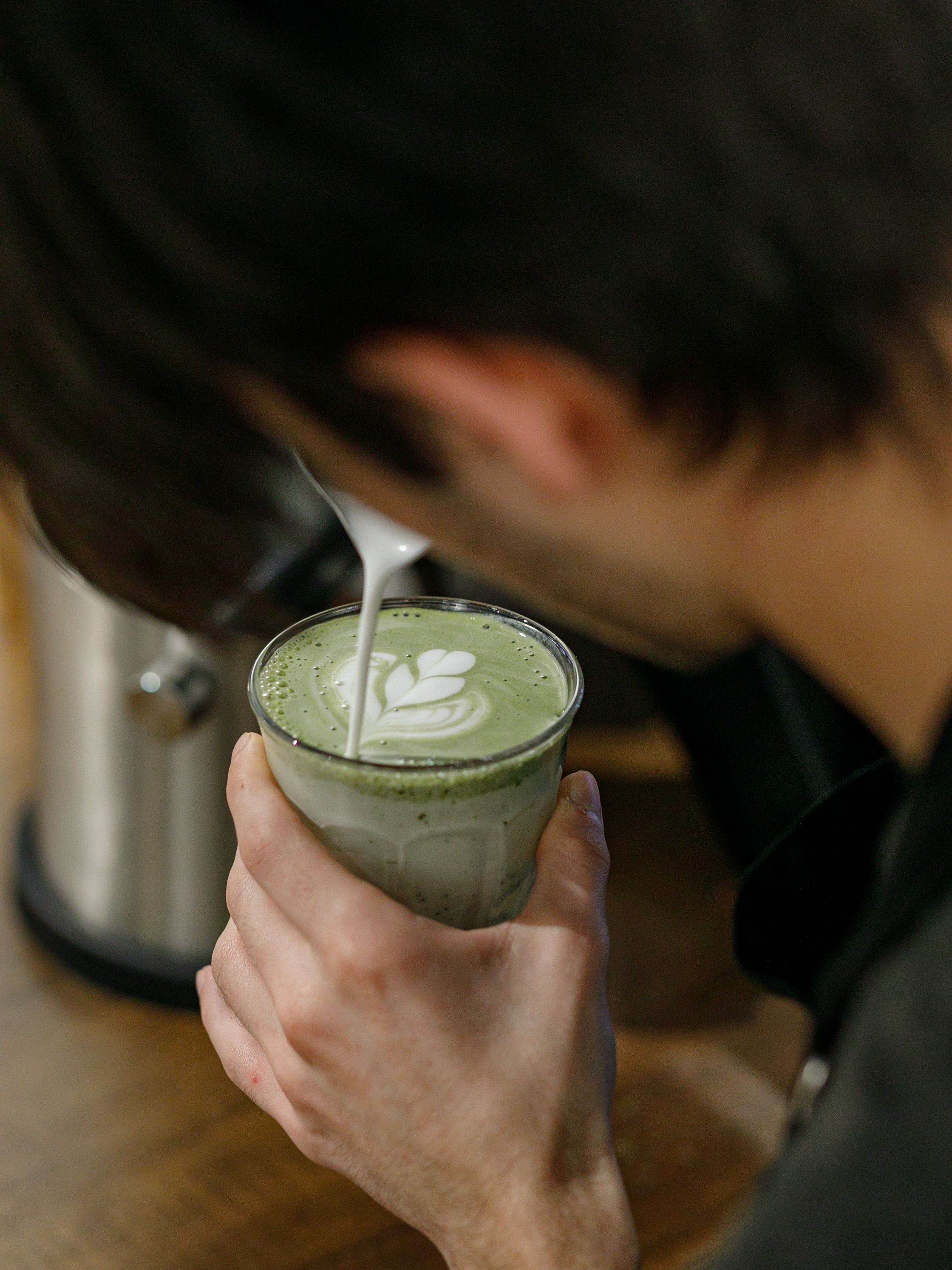 Warm milk being poured into a green tea latte held in a clear glass