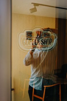 A photographer's reflection in a coffee shop window displaying a decorative logo.