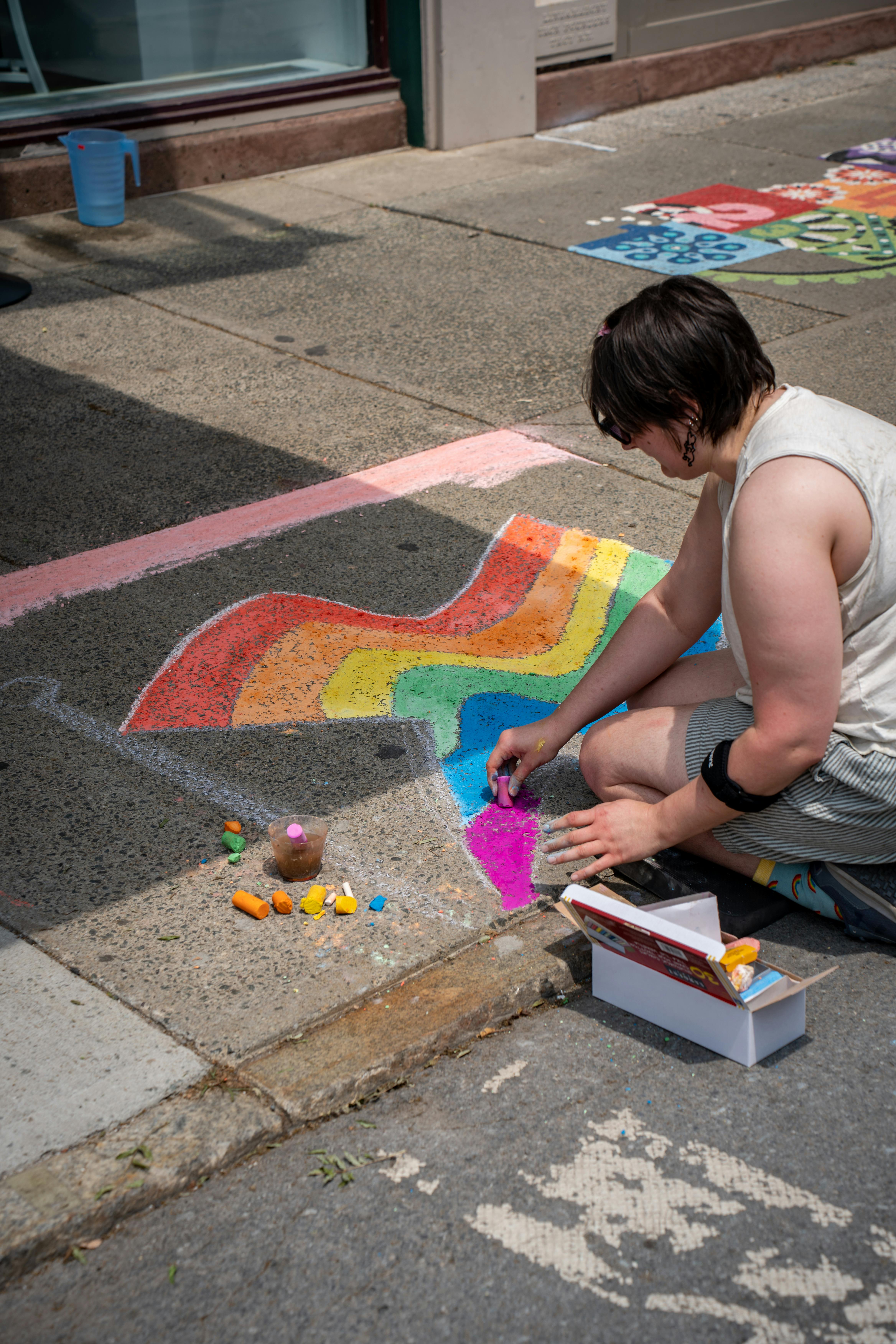 A person drawing vibrant rainbow chalk art on a city sidewalk during the day.