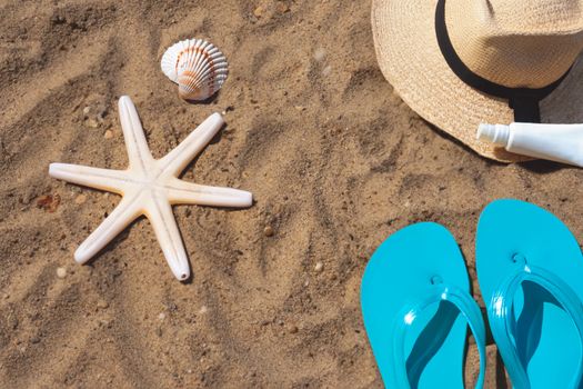 Flat lay of beach essentials including flip flops, hat, starfish, and shells on sand.