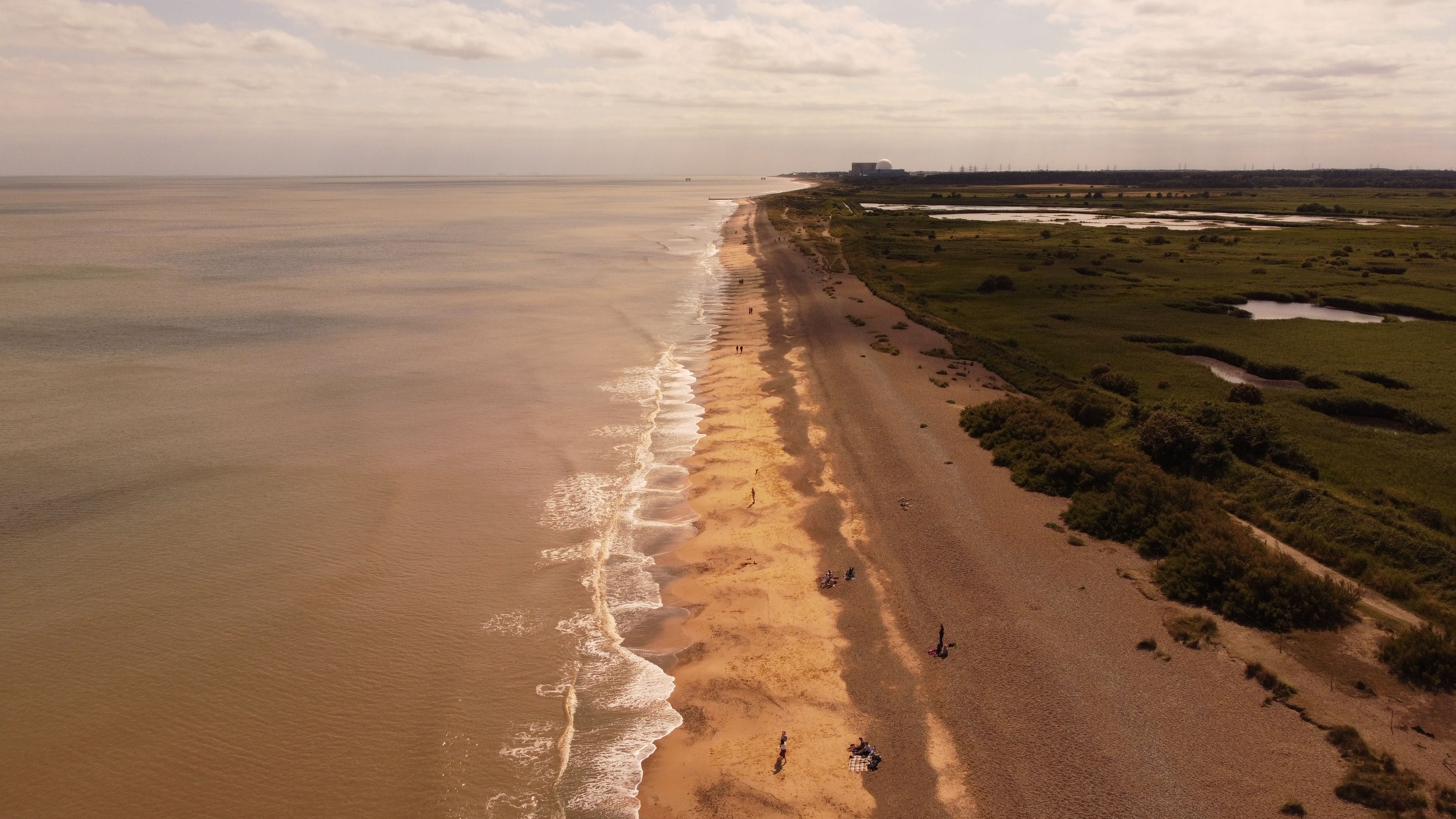A sweeping aerial view of a tranquil coastline with golden sand meeting the expansive ocean under a bright day sky.
