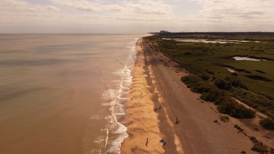 A sweeping aerial view of a tranquil coastline with golden sand meeting the expansive ocean under a bright day sky.