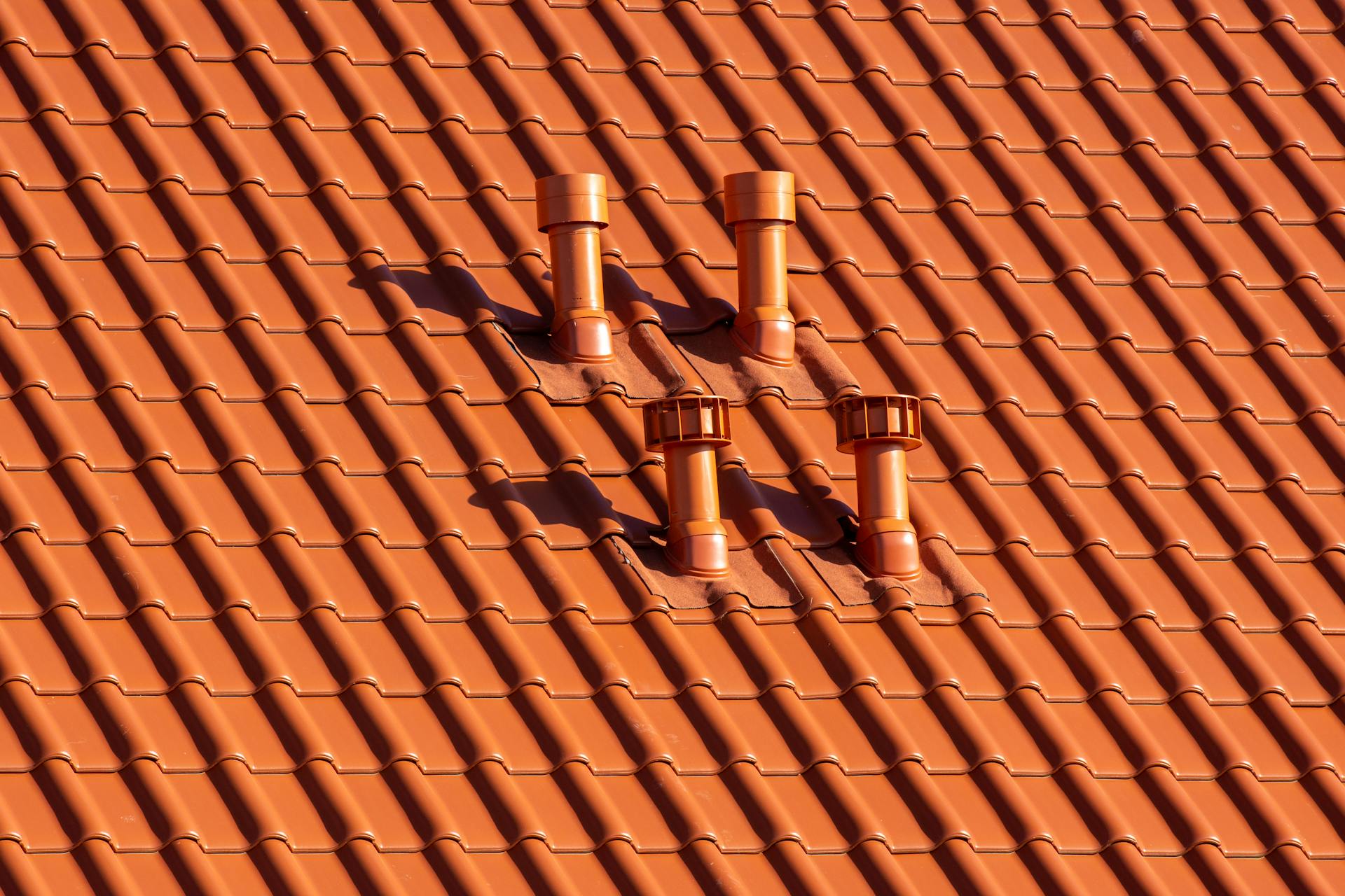 Close-up of an orange tiled roof with chimneys casting shadows in bright sunlight.